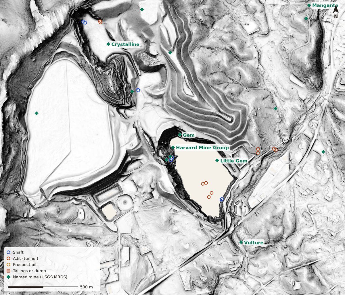 Bare-earth lidar relief of the Jamestown Mine with mine markers: a blank lake-shaped hole where the open pit has filled with water, stepped waste-rock dumps with switchback haul roads to the north, and a large smooth flat oval to the west where the capped tailings facility is