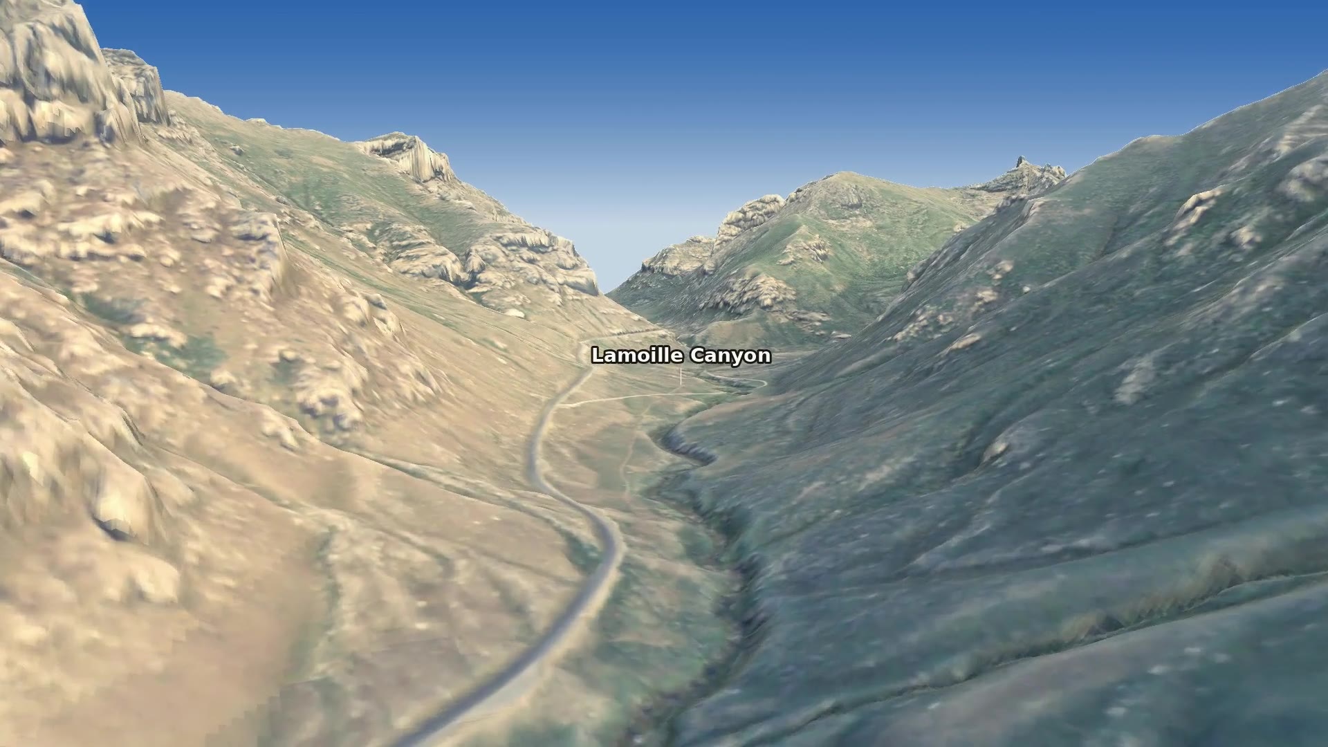 Flying low up Lamoille Canyon: the road and creek wind along a broad, flat floor between steep walls, the left wall warm in late sun and the right wall in shade